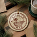 A circular ornament with the text "SALALAH OMAN" printed on it, surrounded by pine branches and a cup of coffee on a wooden surface.
