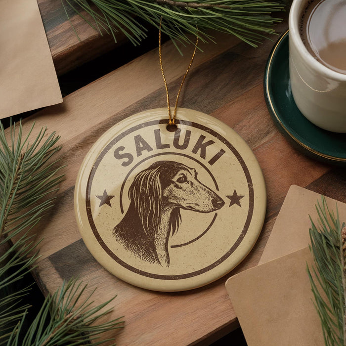 A round ornament with the name "Saluki" and a dog's head image is displayed on a wooden surface, surrounded by pine branches and a cup of coffee.