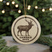 A ceramic ornament featuring a deer with antlers, with the text "SAMBAR DEER" printed on it, is displayed in front of a Christmas tree with twinkling lights.