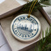 A decorative ornament in a box, featuring a circular design with the text "SAN DIEGO CALIFORNIA" and an illustration of the San Diego skyline.