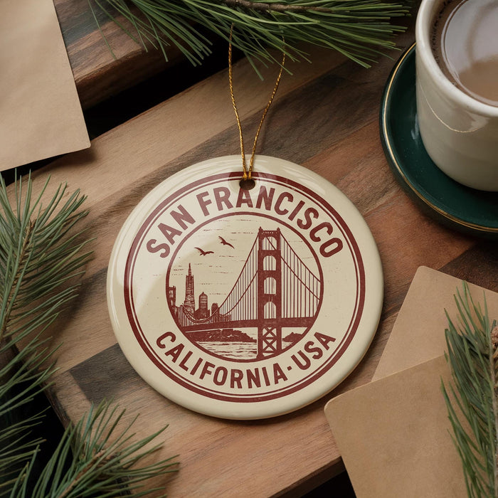 A circular ornament with the text "SAN FRANCISCO CALIFORNIA, USA" and a graphic of the Golden Gate Bridge, placed on a wooden surface alongside a cup of coffee and pine branches.