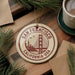 A circular ornament with the text "SAN FRANCISCO CALIFORNIA, USA" and a graphic of the Golden Gate Bridge, placed on a wooden surface alongside a cup of coffee and pine branches.