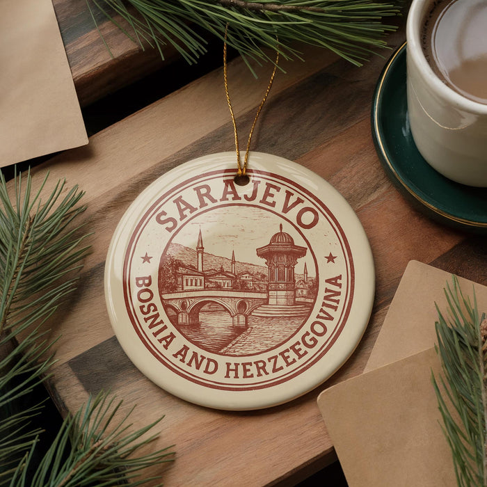 A circular ornament with the text "SARajevo Bosnia and Herzegovina" and an illustration of a bridge is displayed on a wooden surface, accompanied by pine branches and a cup of coffee.