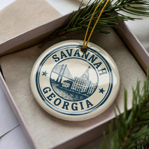 A decorative ornament in a box, featuring a circular design with the text "SAVANNAH GEORGIA" and an illustration of a bridge.