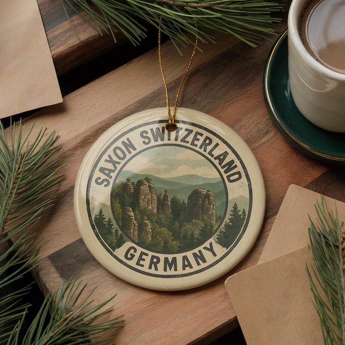 A round ornament with the text "SAXON SWITZERLAND GERMANY" and a scenic mountain landscape is displayed on a wooden surface, accompanied by pine branches and a cup of coffee.