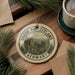 A round ornament with the text "SAXON SWITZERLAND GERMANY" and a scenic mountain landscape is displayed on a wooden surface, accompanied by pine branches and a cup of coffee.