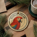 A ceramic ornament featuring a red parrot is displayed on a wooden surface, accompanied by a cup of coffee and some pine branches.