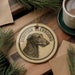 A circular ornament featuring a Scottish Deerhound dog's head, with the text "SCOTTISH DEERHOUND" printed on it, is displayed on a wooden surface alongside a cup of coffee and some pine branches.