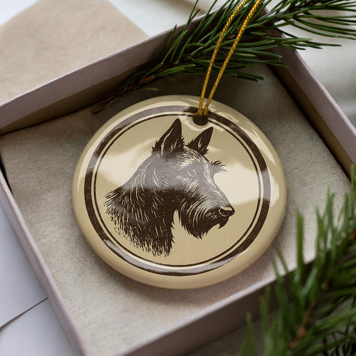 A circular ornament with a black and white illustration of a dog's head is displayed in a box, surrounded by pine branches.