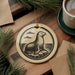 A round ornament featuring a seal sitting on a rock, surrounded by pine branches and a cup of coffee on a wooden surface.