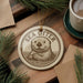 A decorative ornament featuring a sea otter is displayed on a wooden surface, accompanied by a cup of coffee and pine branches.