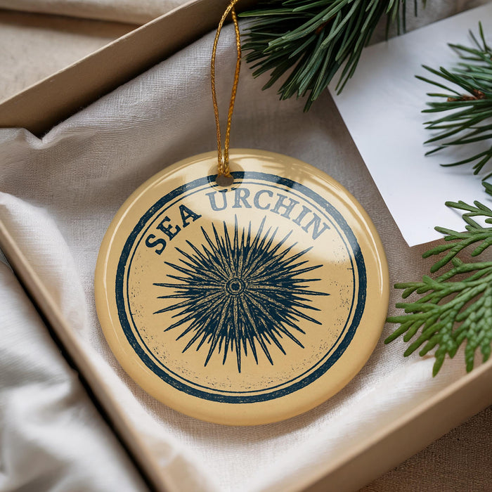 A decorative ornament with the text "SEA URCHIN" on it, hanging from a string. The ornament is placed in a box with pine branches and a white paper.
