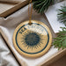 A decorative ornament with the text "SEA URCHIN" on it, hanging from a string. The ornament is placed in a box with pine branches and a white paper.