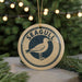 A round ornament with the word "SEAGULL" and a graphic of a seagull is displayed on a table, surrounded by pine branches and twinkling lights.