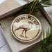 A round ornament with the text "SECRETARY BIRD" and an illustration of a bird is displayed in a box, surrounded by pine branches.