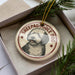 A circular ornament featuring a dog's face, with the text "SHEPADOOD" and "DOG" printed on it, is displayed in a box surrounded by pine branches.