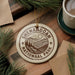 A circular ornament with the text "Shenandoah National Park" and a mountain landscape design hangs on a wooden surface, surrounded by pine branches and a cup of coffee.