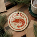 A round ornament with the word "SHRIMP" in red text, featuring a red shrimp illustration, is displayed on a wooden surface alongside a cup of coffee and some pine branches.