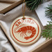 A round ornament with a shrimp design is placed inside a box, surrounded by pine branches.