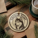 A round ornament featuring a black and white illustration of a Siberian husky dog, with the text "SIBERIAN HUSKY" printed on it. The ornament is hanging from a string, and there is a cup of coffee and some pine branches in the background.
