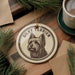 A ceramic ornament featuring a portrait of a silky terrier dog, with the text "SILKY TERRIER" around the image. The ornament is placed on a wooden surface, with pine branches and a cup of coffee nearby.