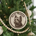 A round ornament with a silhouette of a terrier dog is hanging on a Christmas tree, surrounded by other ornaments and lights.