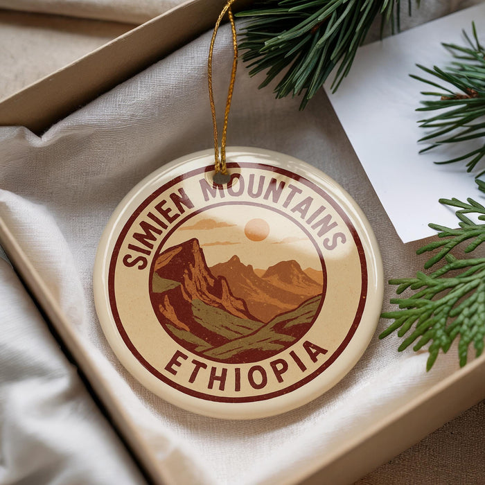 A decorative ornament with the text "SIMIEN MOUNTAINS ETHIOPIA" on it, hanging from a string. The ornament is placed in a box with pine branches and a white paper.