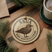 A circular ornament with the text "SKUA BIRD AUSTRALIBIRD" and an illustration of a bird is displayed on a wooden surface, surrounded by pine branches and a cup of coffee.