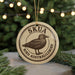 A circular ornament with the text "SKUA BIRD AUSTRALIALDBIRD" and an illustration of a bird, placed on a surface with pine branches in the background.