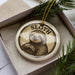 A circular ornament featuring a sloth, a type of slow-moving mammal, is displayed in a box with pine branches.