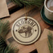 A circular ornament with the name "SLOUGHI" and a dog's head image is displayed on a wooden surface, surrounded by pine branches and a cup of coffee.