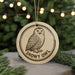 A round ornament featuring a snowy owl, with the text "SNOWY OWL" printed on it, is hanging from a string in front of a Christmas tree.