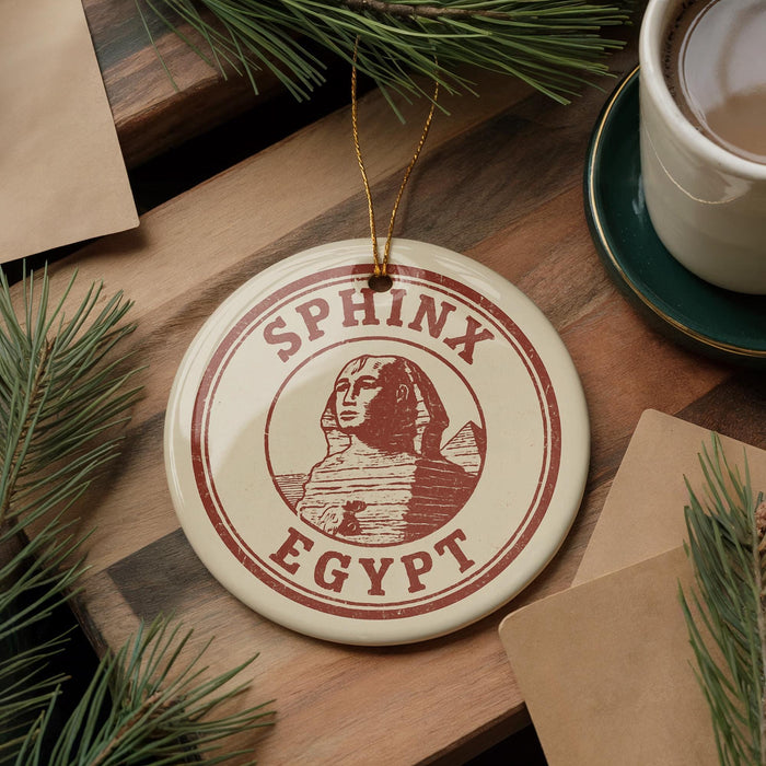A circular ornament with the text "SPHINX EGYPT" and an illustration of the Sphinx is displayed on a wooden surface, surrounded by pine branches and a cup of coffee.