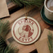 A circular ornament with the text "SPHINX EGYPT" and an illustration of the Sphinx is displayed on a wooden surface, surrounded by pine branches and a cup of coffee.