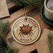 a round ornament with a spider design, placed on a wooden surface next to a cup of coffee and some pine branches.