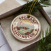 A decorative ornament in a box, featuring a circular design with a scenic image of Spokane, Washington.