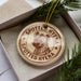 A circular ornament with a spotted hyena image and the text "SPOTTED HYENA" printed on it, placed inside a box with pine branches.