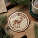 A round ornament with the word "Springbok" printed on it, featuring an illustration of a gazelle or deer. The ornament is hanging from a string, and there is a cup of coffee and some pine branches in the background.