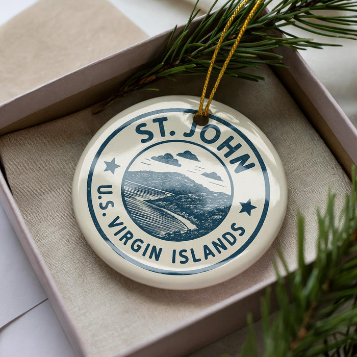 A white ornament with a blue design, featuring the text "ST. JOHN U.S. VIRGIN ISLANDS" and a landscape illustration, is displayed in a box with pine branches.