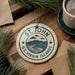 A circular ornament with the text "ST. JOHN U.S. VIRGIN ISLANDS" is displayed on a wooden surface, surrounded by pine branches and a cup of coffee.