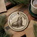 A round ornament featuring a seal on a wooden surface, with a cup of coffee and pine branches in the background.