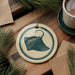 a round ornament with a blue and white design, depicting a stingray, placed on a wooden surface alongside a cup of coffee and some pine branches.