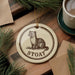 A round ornament featuring a cartoon illustration of a weasel, with the word "STOAAT" written around the edge. The ornament is placed on a wooden surface, surrounded by pine branches and a cup of coffee.