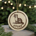 A round ornament with a brown and white illustration of a weasel, and the word "STOAAT" written below it, is hanging from a string.