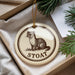 A round ornament with a drawing of a weasel and the word "STOAAT" printed on it, placed in a box with pine branches.