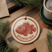A round ornament featuring a red and white depiction of a stonefish is displayed on a wooden surface, surrounded by pine branches and a cup of coffee.