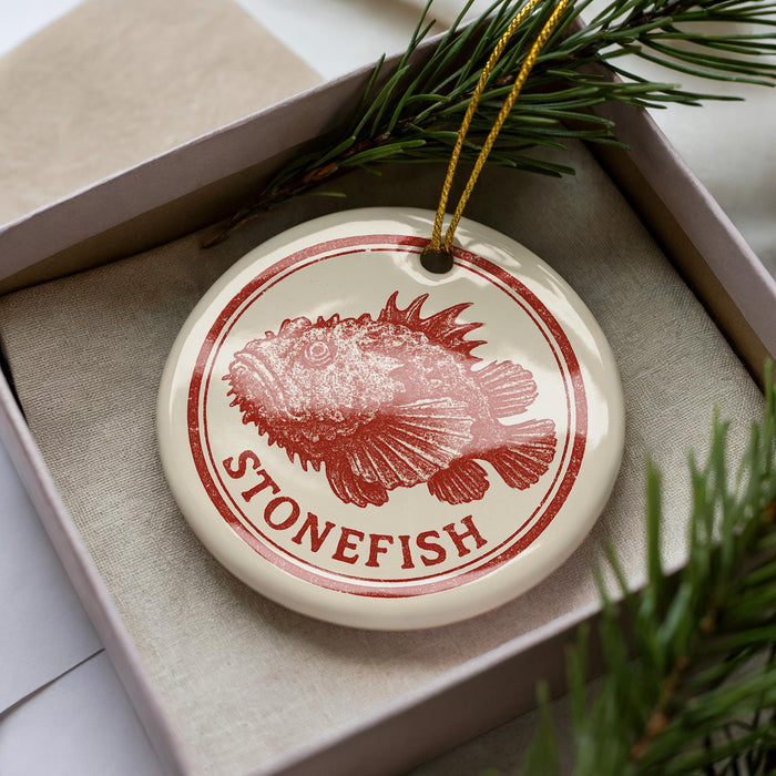 A round ornament with a red and white design featuring a fish, placed inside a box with pine branches.