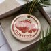 A round ornament with a red and white design featuring a fish, placed inside a box with pine branches.