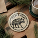 A decorative ornament featuring a striped hyena, with the text "STRIPED HYENA" printed on it, is displayed on a wooden surface alongside a cup of coffee and some pine branches.