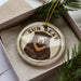 A circular ornament featuring a bear image is displayed in a box, surrounded by pine branches.
